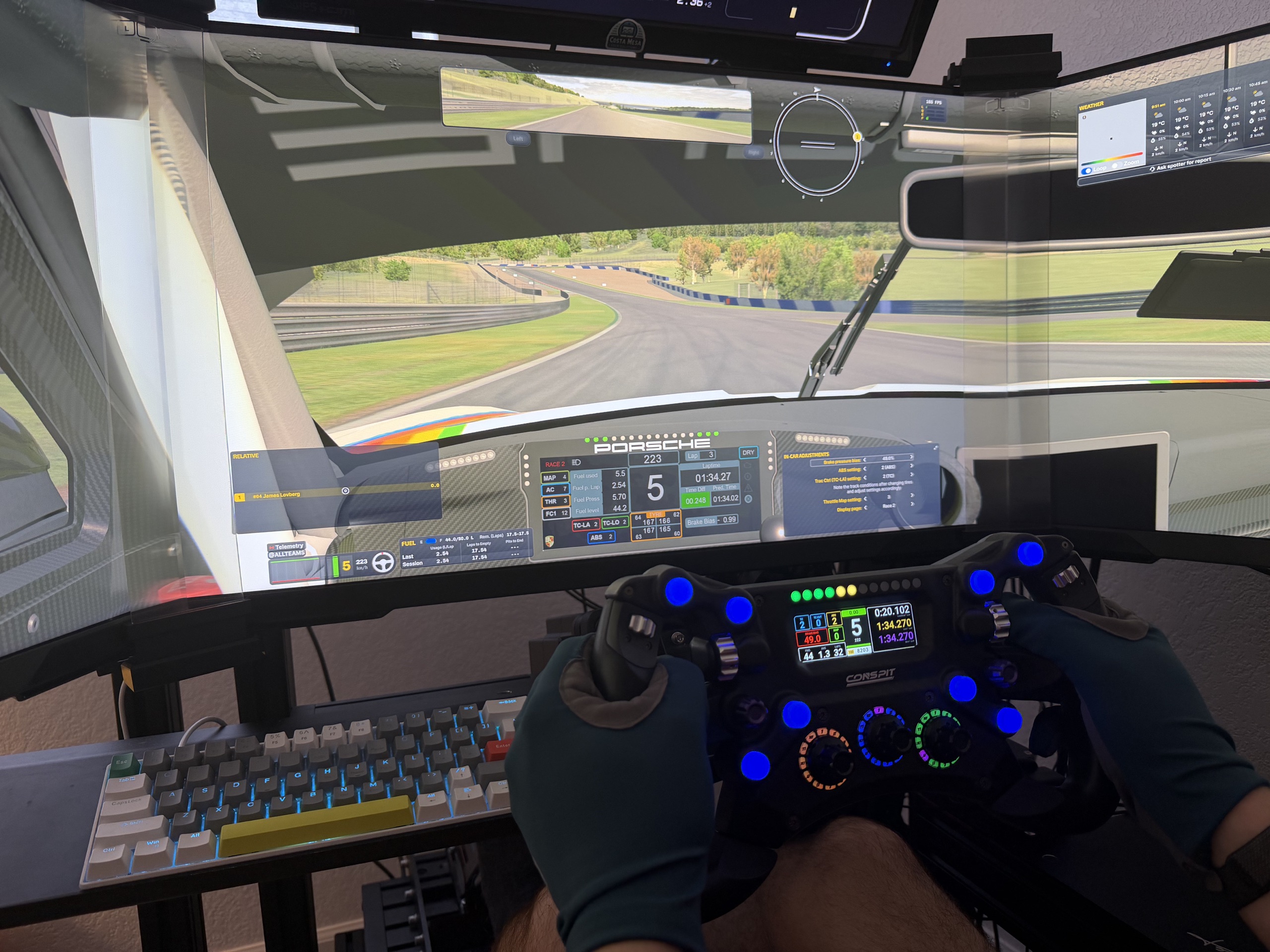 Driver holding a sim racing steering wheel in front of a triple-screen Porsche cockpit.